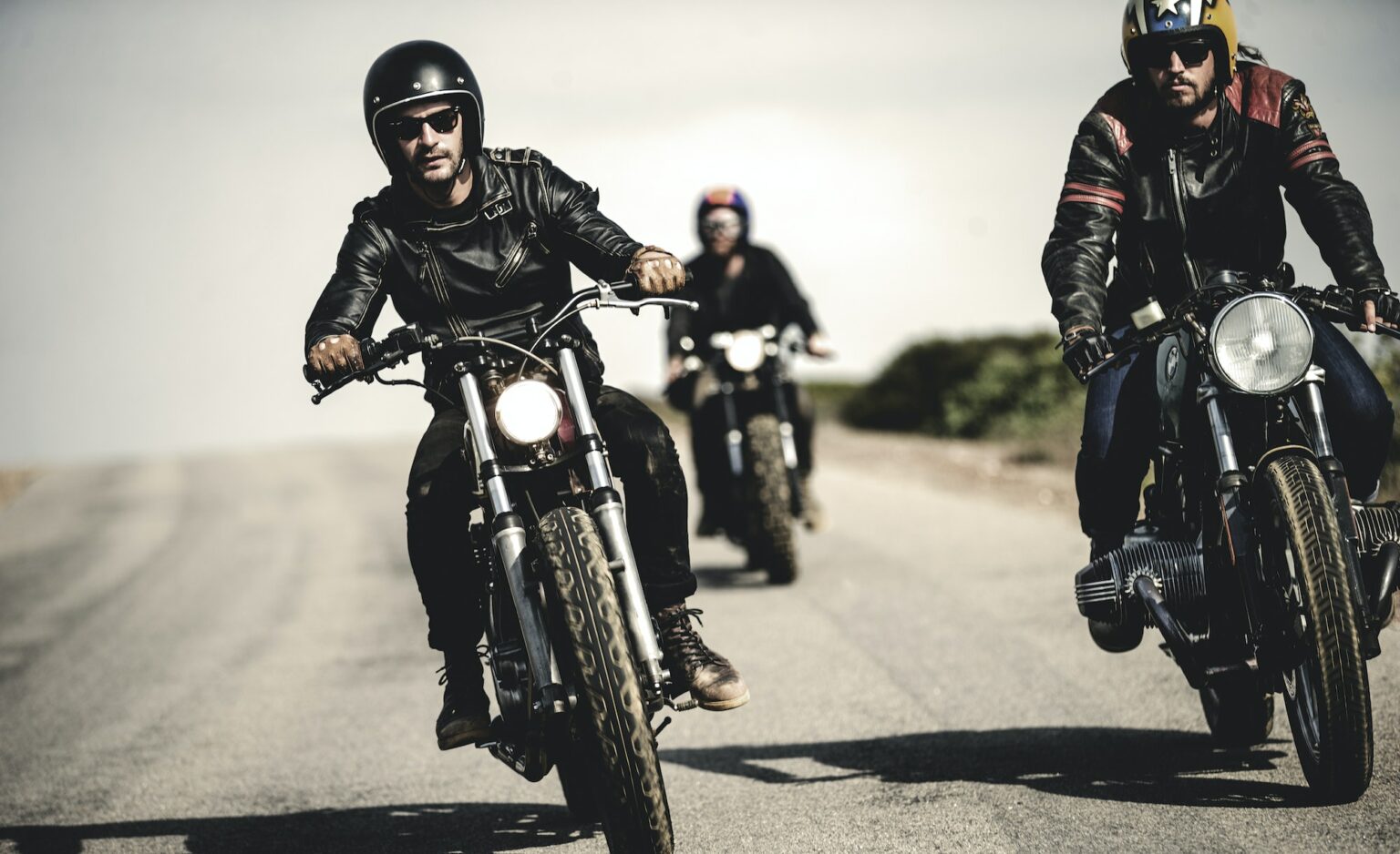 Three men wearing open face crash helmets and sunglasses riding cafe racer motorcycles along rural
